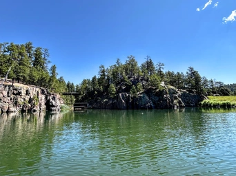 View of the cliffs along the shore of Sheridan Lake in the Black Hills, SD. Blue skies, a bridge, and pine trees.