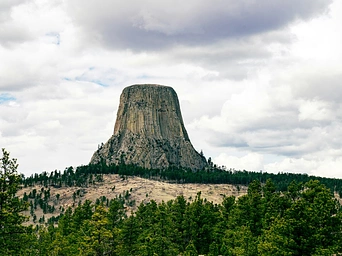 Devils Tower Monument in Wyoming wit pine trees in the foreground and cloudy skies.