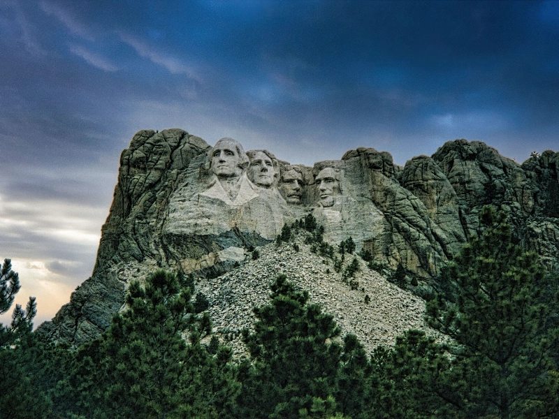 Mount Rushmore in the Black Hills with pine trees in front and clouds overhead.